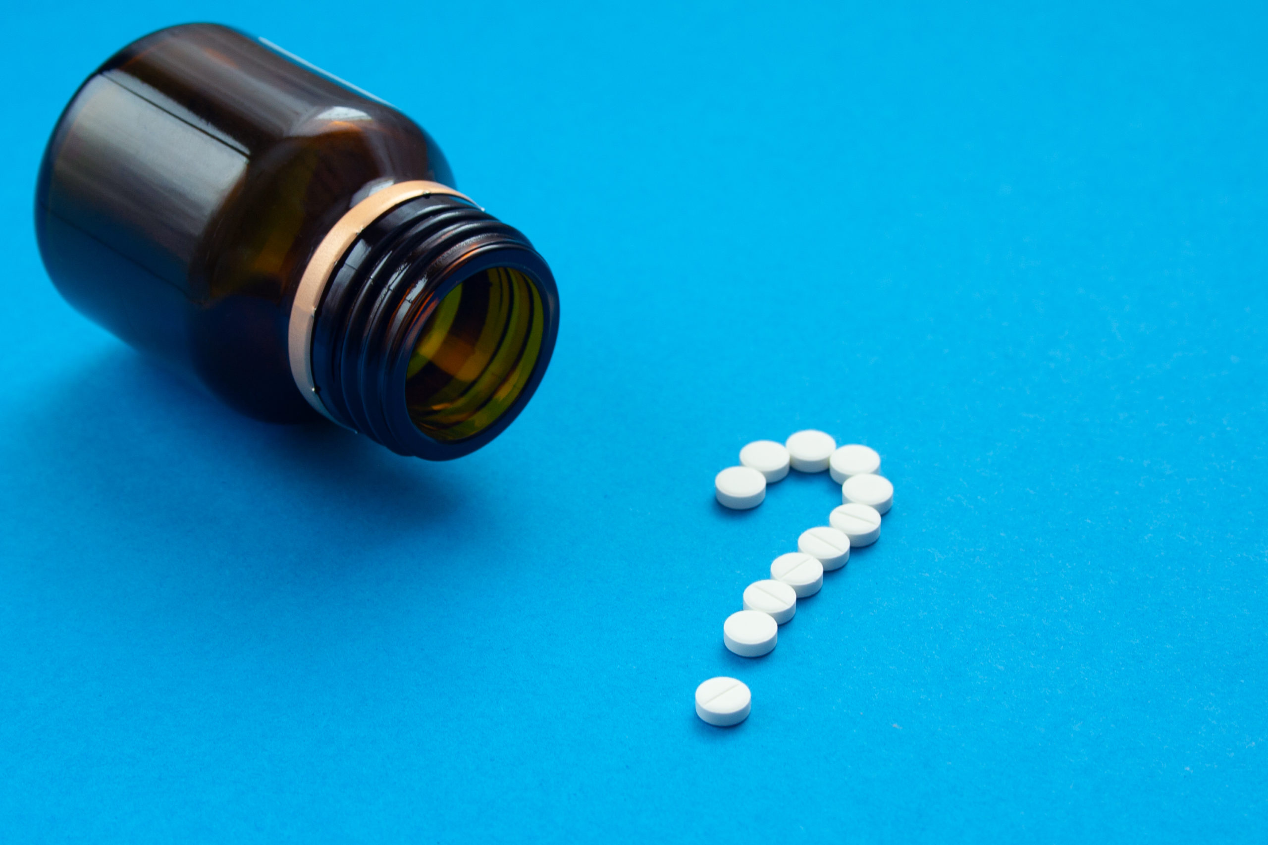white pills in the shape of a question mark are scattered on a blue background next to a dark bottle.